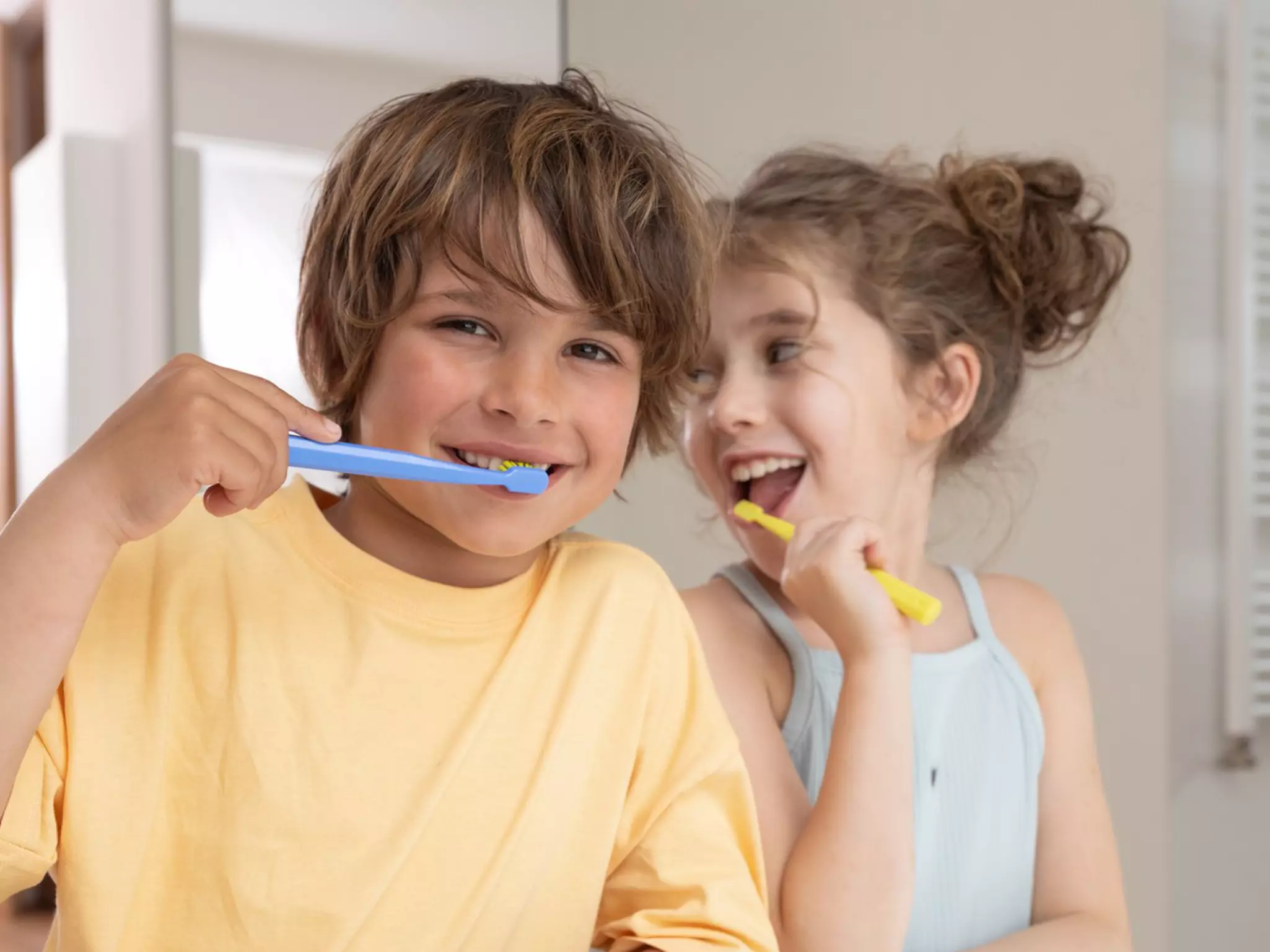 Zwei unterschiedlich alte Kinder stehen nebeneinander im Badezimmer und putzen sich die Zähne, eines mit einer blauen Zahnbürste, das andere mit einer gelben. Das Bild dient als Sinnbild dafür, dass Zahnwechsel bei Kindern unterschiedliche Zahnpflege erfordert.