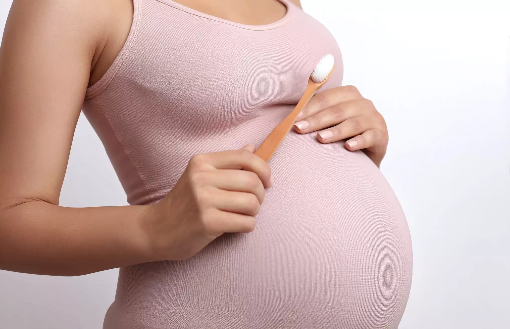 Ausschnitt einer seitlich stehenden Frau von der Brust bis zum Bauch, die sich mit der hinteren Hand den schwangeren Bauch hält und mit der vorderen Hand eine Zahnbürste hält; als Sinnbild für Zahnpflege während der Schwangerschaft.